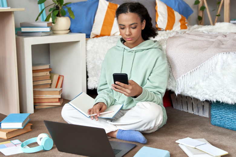 Representing creating experiences with your teen - A teen girl sitting on the floor of her bedroom doing her homework with a laptop on the floor and phone in her hand.