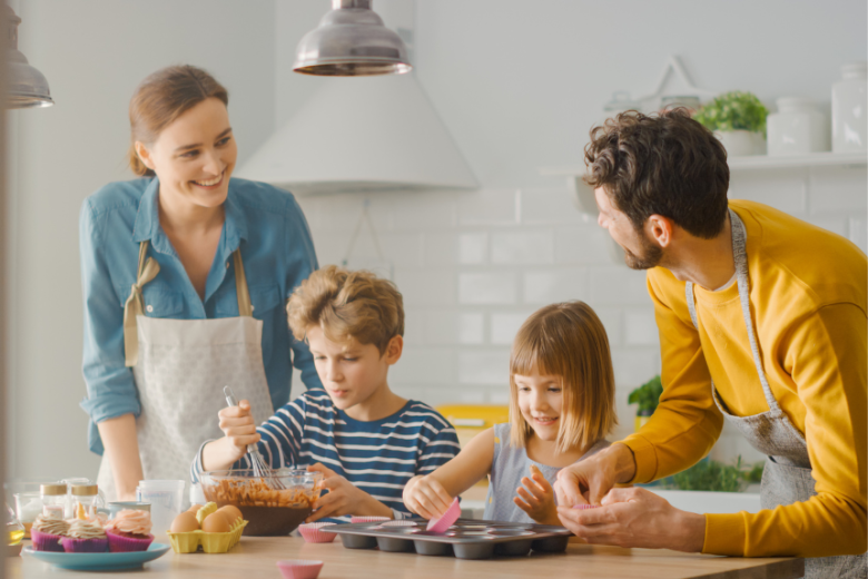 Image representing foster care approval in the North West. A young boy and girl decorating cupcakes on a kitchen bench being supervised by a smiling woman and man who is looking at her, both wearing aprons.